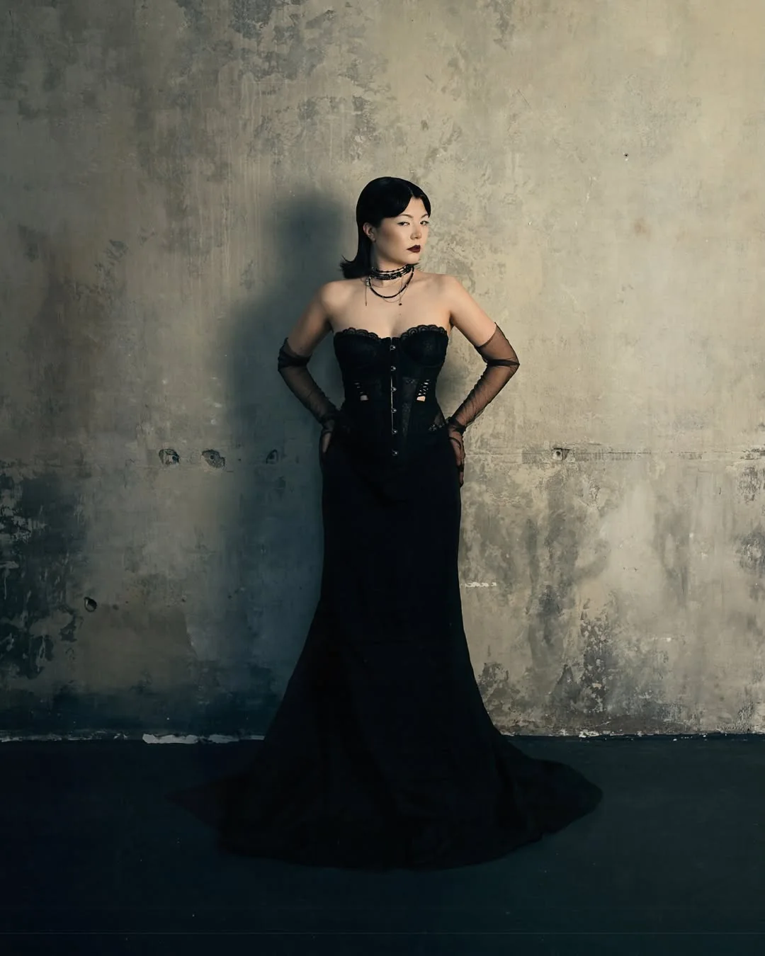 Full-length black corset gown against a concrete wall